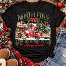 Load image into Gallery viewer, North Pole Christmas Tree Farm vintage holiday graphic shirt featuring a red pickup truck carrying Christmas trees, snowy evergreen trees, rustic farmhouse scenery, festive red bows, and distressed green lettering reading North Pole Christmas Tree Farm Always Fresh. Available in beige and black in short sleeve crewneck, V-neck, long sleeve crewneck, and hoodie styles.