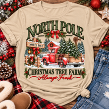 Load image into Gallery viewer, North Pole Christmas Tree Farm vintage holiday graphic shirt featuring a red pickup truck carrying Christmas trees, snowy evergreen trees, rustic farmhouse scenery, festive red bows, and distressed green lettering reading North Pole Christmas Tree Farm Always Fresh. Available in beige and black in short sleeve crewneck, V-neck, long sleeve crewneck, and hoodie styles.