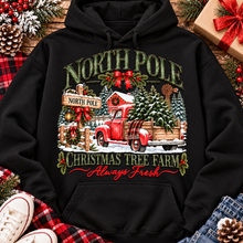 Load image into Gallery viewer, North Pole Christmas Tree Farm vintage holiday graphic shirt featuring a red pickup truck carrying Christmas trees, snowy evergreen trees, rustic farmhouse scenery, festive red bows, and distressed green lettering reading North Pole Christmas Tree Farm Always Fresh. Available in beige and black in short sleeve crewneck, V-neck, long sleeve crewneck, and hoodie styles.