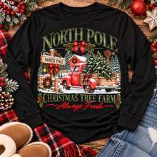 Load image into Gallery viewer, North Pole Christmas Tree Farm vintage holiday graphic shirt featuring a red pickup truck carrying Christmas trees, snowy evergreen trees, rustic farmhouse scenery, festive red bows, and distressed green lettering reading North Pole Christmas Tree Farm Always Fresh. Available in beige and black in short sleeve crewneck, V-neck, long sleeve crewneck, and hoodie styles.