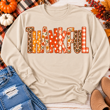 Load image into Gallery viewer, Beige long sleeve crewneck shirt featuring colorful Thankful patchwork lettering in retro autumn floral, plaid and polka dot patterns.
