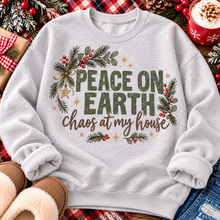 Load image into Gallery viewer, Peace on Earth Chaos at My House funny Christmas graphic apparel featuring distressed olive green lettering, rustic pine greenery, red berries and gold stars, available in sand and sport grey in short sleeve crewneck tees, V-neck tees, sweatshirts and hoodies.
